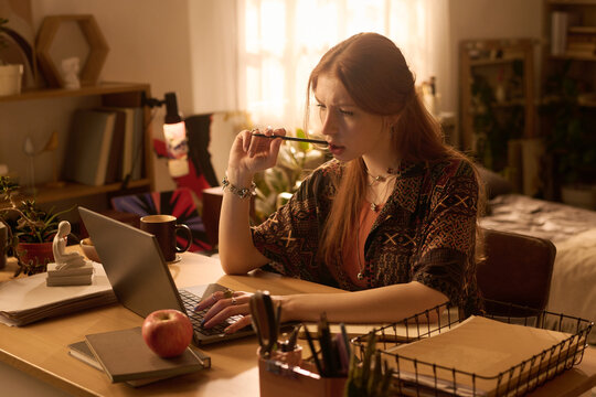 Young adult Caucasian woman sitting at desk working on laptop, holding pencil to mouth, appearing focused, surrounded by books and stationery in home office or study environment - Powered by Adobe
