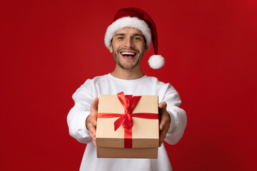 A man wearing a Santa hat presents a festive gift box with a red ribbon. He is smiling widely,...
