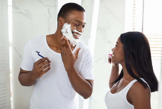 A man is smiling as he shaves his face while a woman watches and applies makeup in a bright, stylish bathroom. They share a light moment during their morning routine. - Powered by Adobe