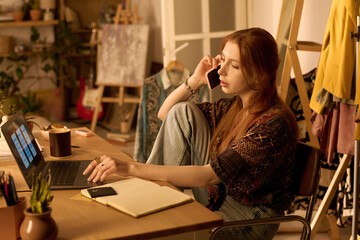 Young adult Caucasian woman sitting at desk working on laptop, holding phone to ear, surrounded by notebook and coffee cup, appearing focused in creative workspace