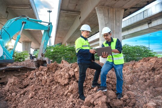 Construction Workers Collaborating on Project Progress Under Overpass with Excavator and Loamy Soil