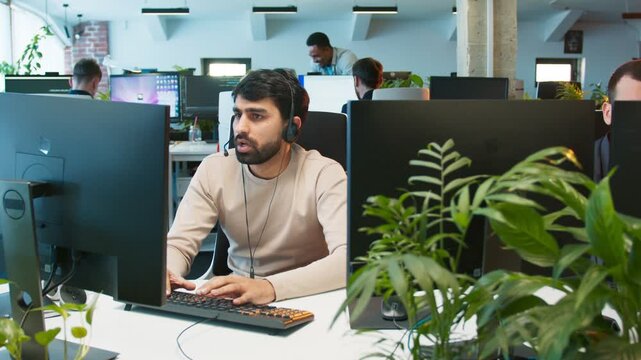 Male wearing headset assisting client. Man talking while checking data on monitor. Typing while solving issue for customer. Colleagues working at computers in bright office with plants around.