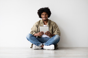 A young African American man sits comfortably on the floor indoors. He is smiling while using a digital tablet for browsing the internet. The setting is bright and simple with a white wall behind him.