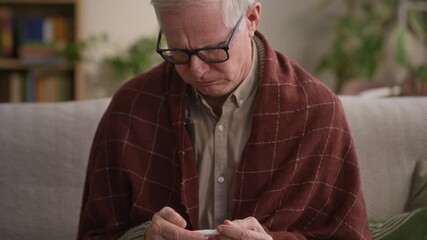 Tilt up shot of mature Caucasian man with flu measuring temperature with thermometer while sitting on sofa wrapped in blanked at home