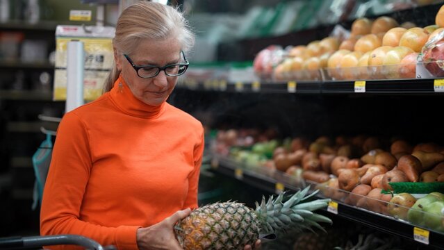 Mature woman in orange sweater looks closely at a pineapple while shopping in a grocery store filled with fresh fruits and vegetables - Powered by Adobe
