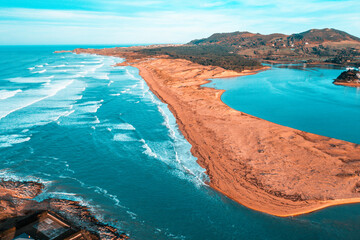 Drone shot of the Liencres coastline, revealing waves crashing along the sandbar while the serene blue estuary stretches inland under a clear sky.