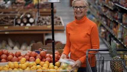 Mature woman smiles while selecting fresh fruits in a grocery store looking for the best deals on food items.