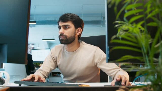 Close camera view of office worker rapidly typing. Male submitting work late while focusing on screen. People working in background at desks. Active office environment showing intense workflow.