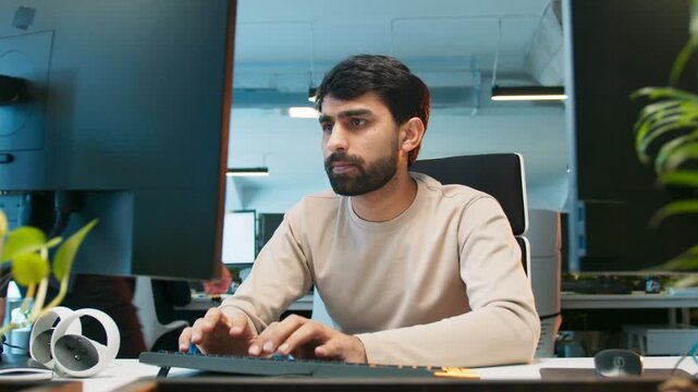 Male with worried look fixing code fast. Programmer busy typing while focusing on screen. People working at desks in background. Active office environment showing intense development workflow.