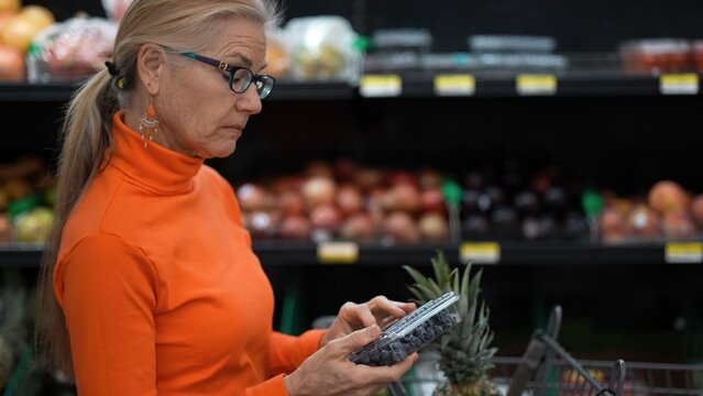 Mature woman examines a container of blueberries while shopping for groceries in a store, focusing on fruits and vegetables.