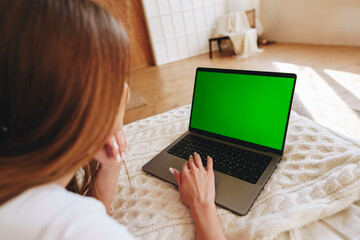 Woman focused on green screen laptop while sitting on bed in cozy room during the day