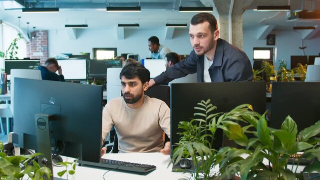 People writing code together. Male receiving help from coworker while focusing on screen. Team working at computers in shared office. Active workspace showing collaborative development process.
