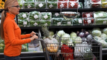 Woman walks through a grocery store aisle pushing a cart filled with various food items while looking at prices to compare.