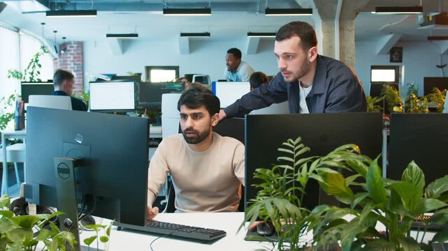 Coworker assisting intern male writing code. Man focusing on keyboard while teammate pointing at monitor. People working at computers in shared office. Collaborative development process.