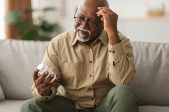 An older man sits on a couch, displaying concern as he examines a bottle of medication. The setting is cozy, illuminated by soft evening light, hinting at his contemplation.