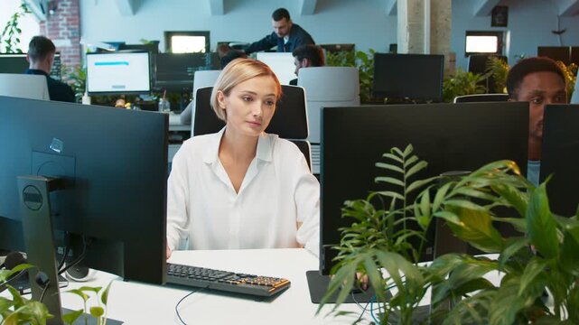 Charming female working on two monitors while creating application. Splitting screens for productivity. People working at computers in background. Using mouse and keyboard while coding.