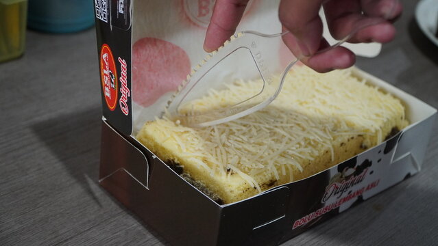Wonosobo, Indonesia - 19 November 2025: Hand slicing traditional cake topped with grated cheese in packaging box.