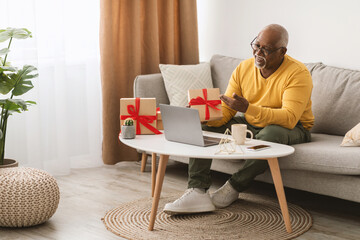A senior man in a yellow sweater enjoys a video call from his cozy living room, surrounded by gifts and indoor plants. He is happily engaging with loved ones.