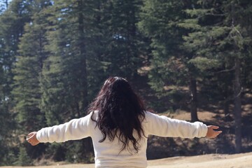 Woman standing with open arms facing tall pine trees in Shimla, expressing peace, adventure, and connection to nature.