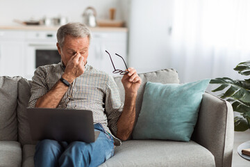 A senior man is seated on a sofa, focused on his laptop. He rubs his eyes in frustration, indicating a challenging moment. The cozy living room has natural light.