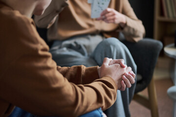 Young man sitting with hands clasped, listening attentively to adult woman holding cards during group therapy session, partial body view