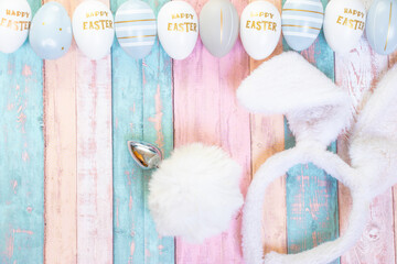 Colorful Easter eggs, some with text "Happy Easter" and anal plug like bunny tail and ears decorated on pastel wood as a background.