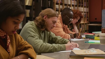 Side view of focused male student writing progress check test while sitting in row with multi ethnic classmates studying in language training course for adults in library