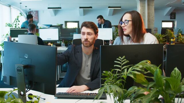 Caucasian female colleague standing beside desk pointing at monitor showing mistake in code. Male focusing on screen while receiving help. Modern office workspace with bright lights and many coworkers