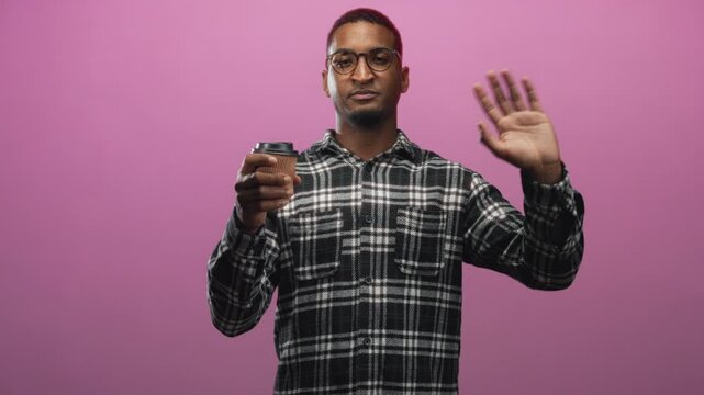 Man wearing glasses and plaid shirt holding paper coffee cup and waving hand in studio with pink backdrop; friendly greeting.