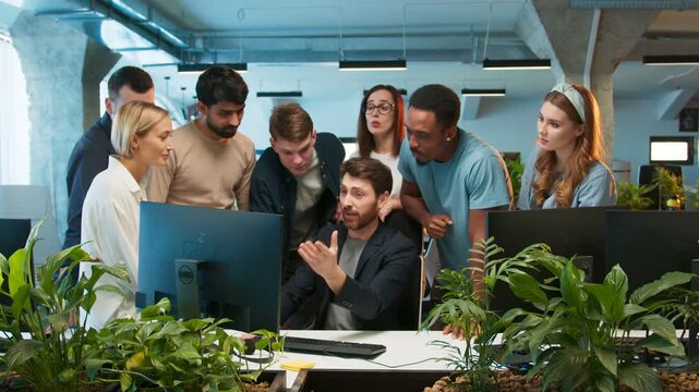 Group of coworkers surrounding male colleague. Team praising his completed task. Smiling man sitting at desk. Open workspace with computers and plants. Green plants located in front.