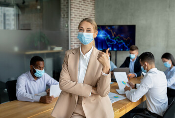 Young business lady poses with a thumbs up in a modern office setting during a corporate meeting. Colleagues work at a table while following health guidelines with face masks.