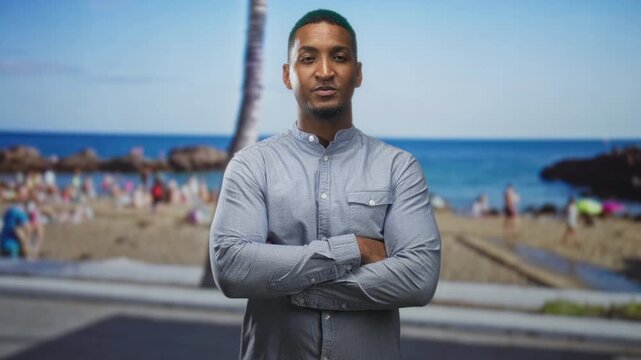 Young black man standing with arms crossed and visible hands on a crowded sandy beach by the sea and palm tree; confidence approachable.