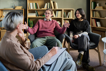 Young man gesturing while speaking during group therapy session with young woman listening and adult woman taking notes on clipboard