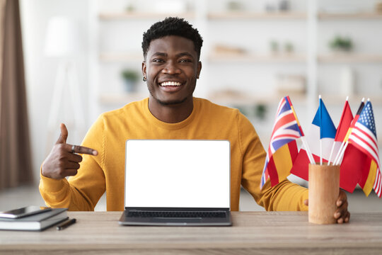 Happy black guy teacher showing language school on laptop with blank screen, mockup, sitting at workdesk with notebook and flags of different countries, office interior, copy space