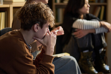 Teenage boy sitting with hand on chin appearing thoughtful while participating in group therapy session with diverse teenagers in background listening attentively