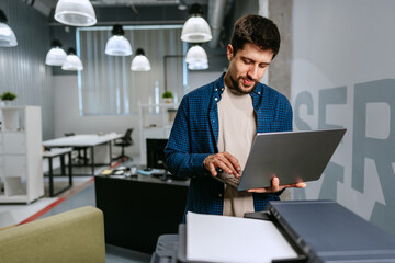 Man working on laptop in modern office setting during daytime while using printer