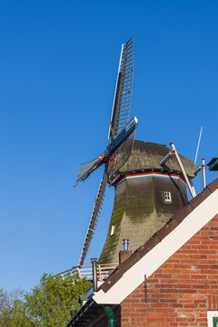 A close-up view of a windmill in Bedum, Netherlands, framed behind a brick building and roof. The prominent blue sky and sharp morning light highlight the structure's details
