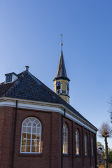 Obraz premium A side view of a brick church with arched windows and a clock tower in Bedum, Netherlands. The building is illuminated by morning sun under a clear blue sky