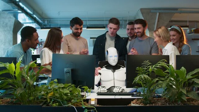 Team of engineers gathered around workstation. Mixed group celebrating robot completing assigned task. Developers smiling as humanoid sits at center. Modern collaborative tech space.