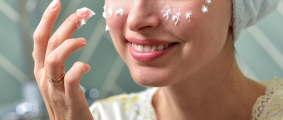 Beautiful Young Woman applying facial moisturizing cream in bathroom.Home interior. Skincare concept