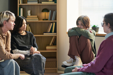 Group of young and adult women and man sitting in circle participating in group therapy session, multiethnic people in conversation, one woman hugging knees