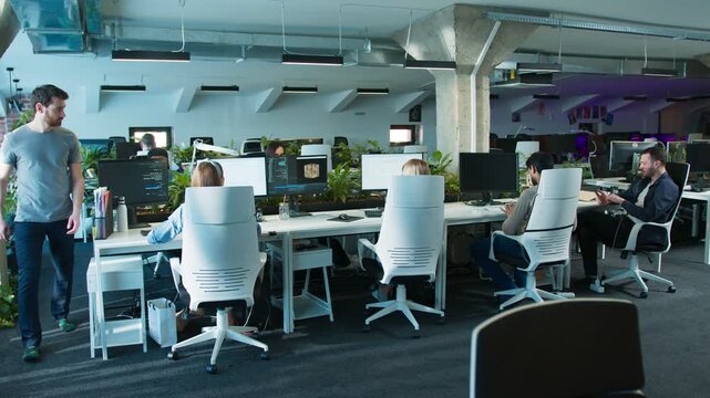 Several coworkers sitting at desks in business center. People working on computers with code on monitors. Male walking beside row of desks. Office area filled with plants and screens.