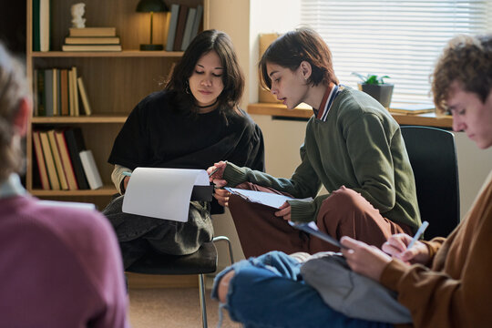 Group of multiethnic teenagers sitting in circle participating in group therapy session, holding papers and pens, discussing and writing notes together in bright room