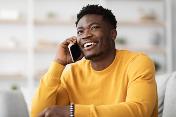 Cheerful african american guy in yellow sitting on sofa with laptop, having phone conversation with employer and smiling, looking at copy space. Handsome millennial black man looking for job