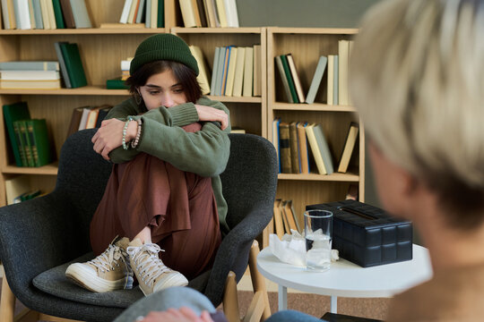 Teenage girl sitting curled up in chair hugging knees during group therapy session, looking down with thoughtful expression while adult woman listening nearby