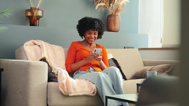 A joyful woman is engaged with her smartphone, smiling as she interacts with her mobile device while sitting on a cozy couch in a welcoming living space, showcasing modern technology and leisure.