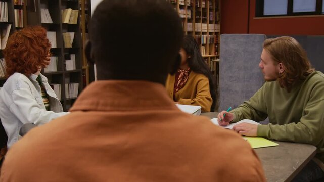 Young South Asian female teacher presenting new grammar rule to multiethnic students in English class pointing at verb tense table on whiteboard while studying in small study group in library