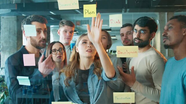 Team placing sticky notes on glass wall. Coworkers focusing on planning task while reviewing ideas. Woman placing note labeled plan as others discuss strategy. Busy project room. Making strategy.