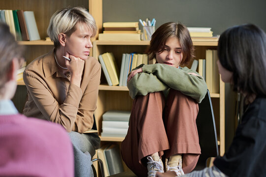 Adult woman facilitating group therapy session with teenage girl sitting with knees to chest and two other young women listening attentively