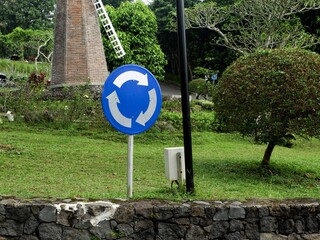 Blue and white circular traffic sign indicating a roundabout or mandatory turn, set in a lush green park with a brick windmill structure in the background. Road sign.